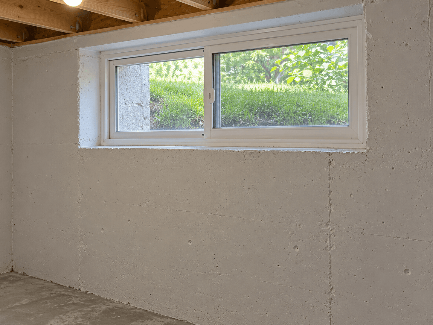A closed high basement window above an exposed foundation wall on a green summer day.