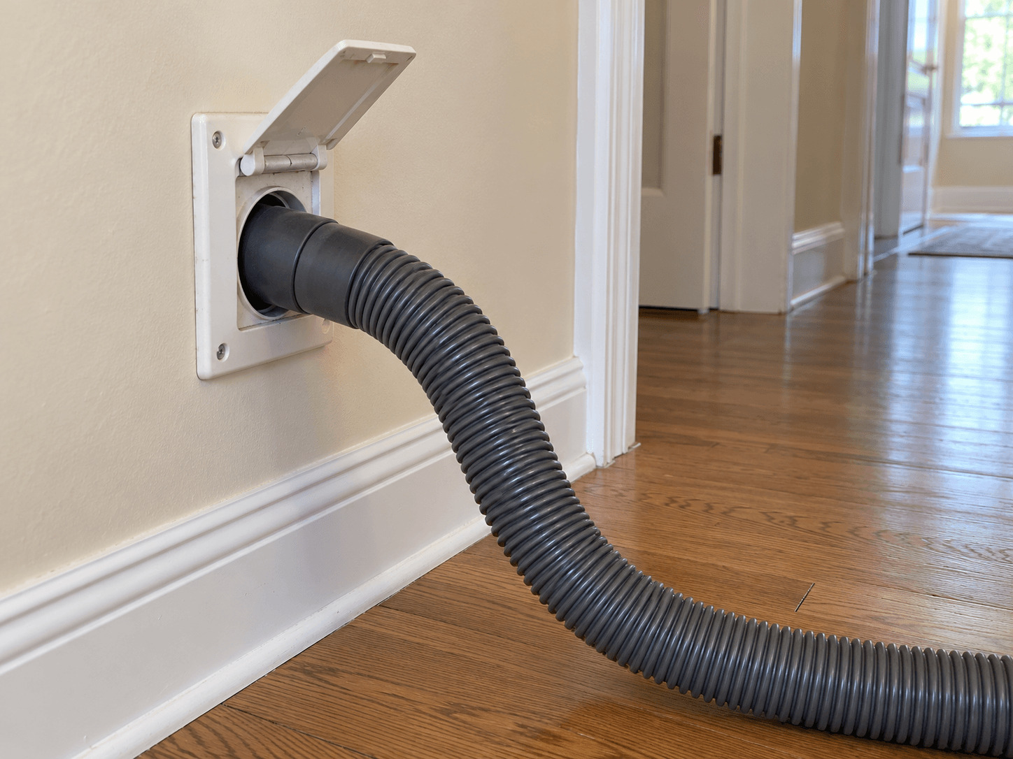 A corrugated central vacuum hose connected to a hinged wall inlet above an oak floor.