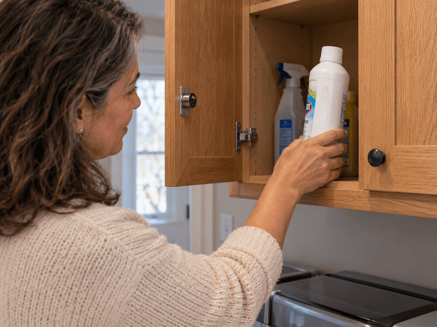 A homeowner returns a capped cleaner in its original bottle to a lockable upper utility cupboard.