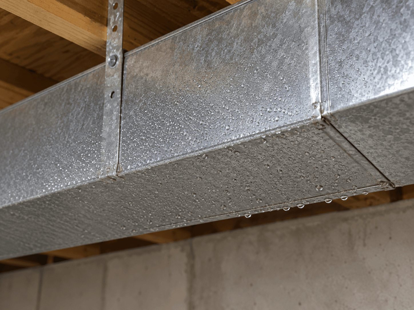 Small droplets collect across the underside of a bare metal cooling duct in an unfinished basement.