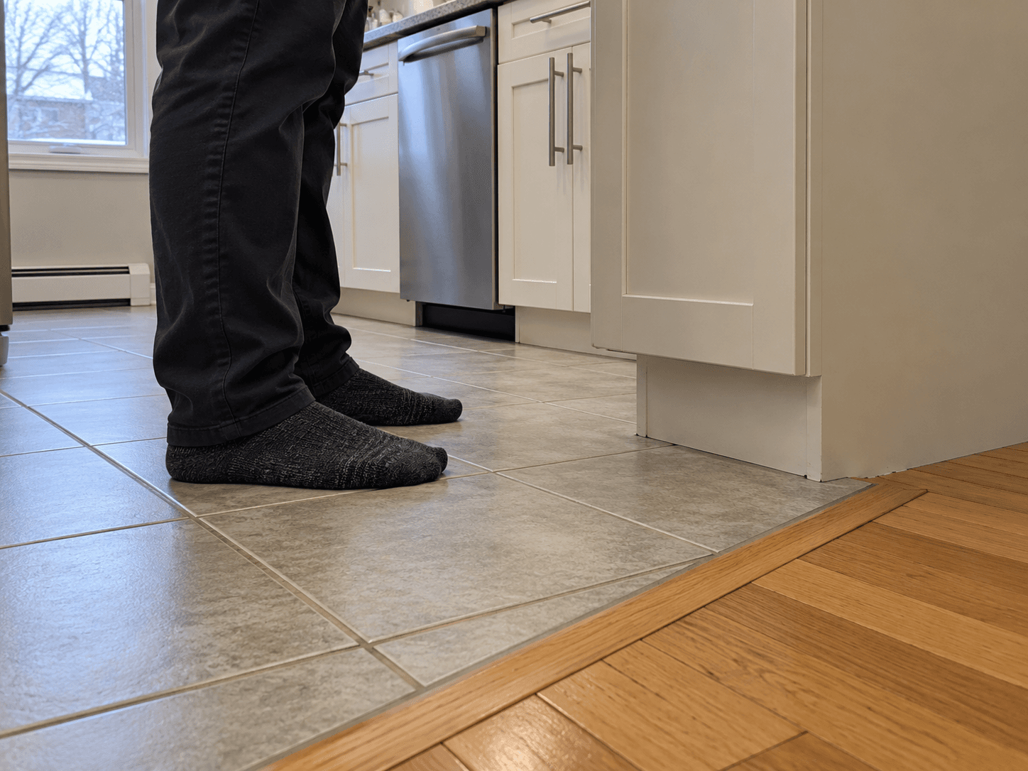 A homeowner's socked feet rest on a tiled kitchen floor beside a clear low wall register.