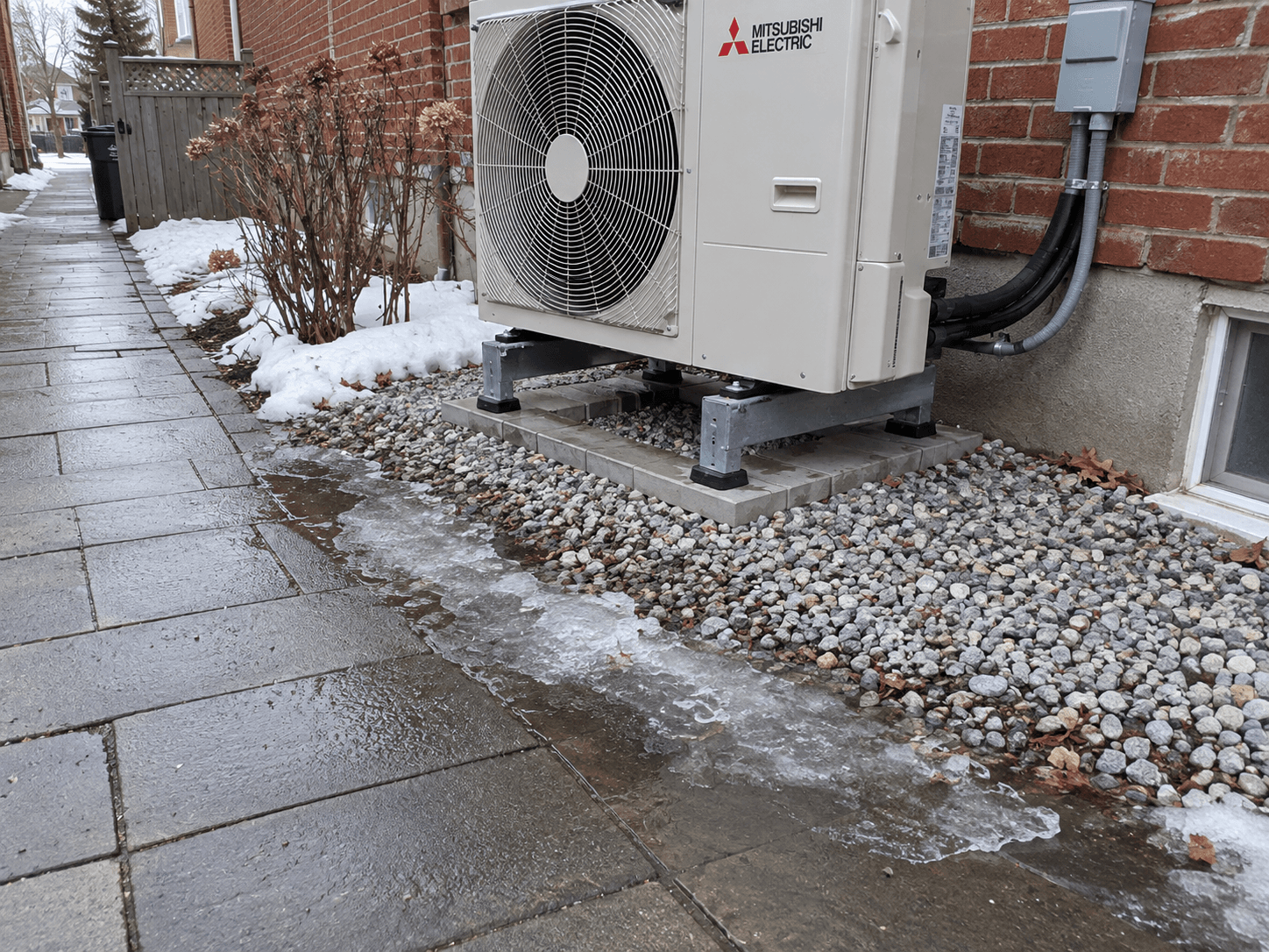 Meltwater and a thin icy patch at the edge of a path beside a raised outdoor heat pump.